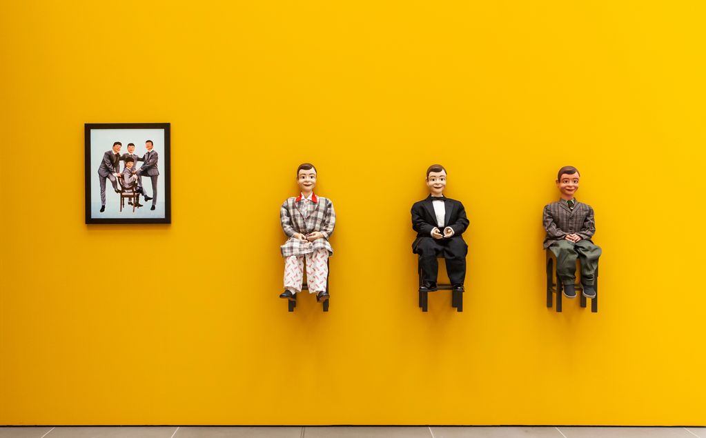 Exhibition view with a photograph of four hand puppets in grey suits, presumably intended to represent the band The Beatles in their younger years. Next to them are three dolls that look the same but wear different suits and have been mounted sitting on the yellow wall. Laurie Simmons, Sammlung Goetz Munich