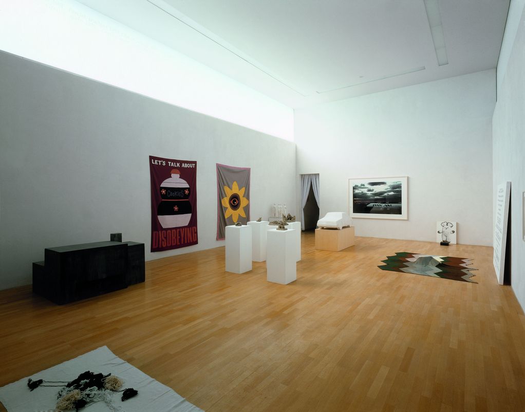Here you can see an exhibition view with works by Mike Kelley and Peter Fischli & David Weiss. In the large, luminous room, two tapestries, several three-dimensional objects on pedestals, two carpets and several two-dimensional works, including a photograph of a Pan Am airplane, can be seen.