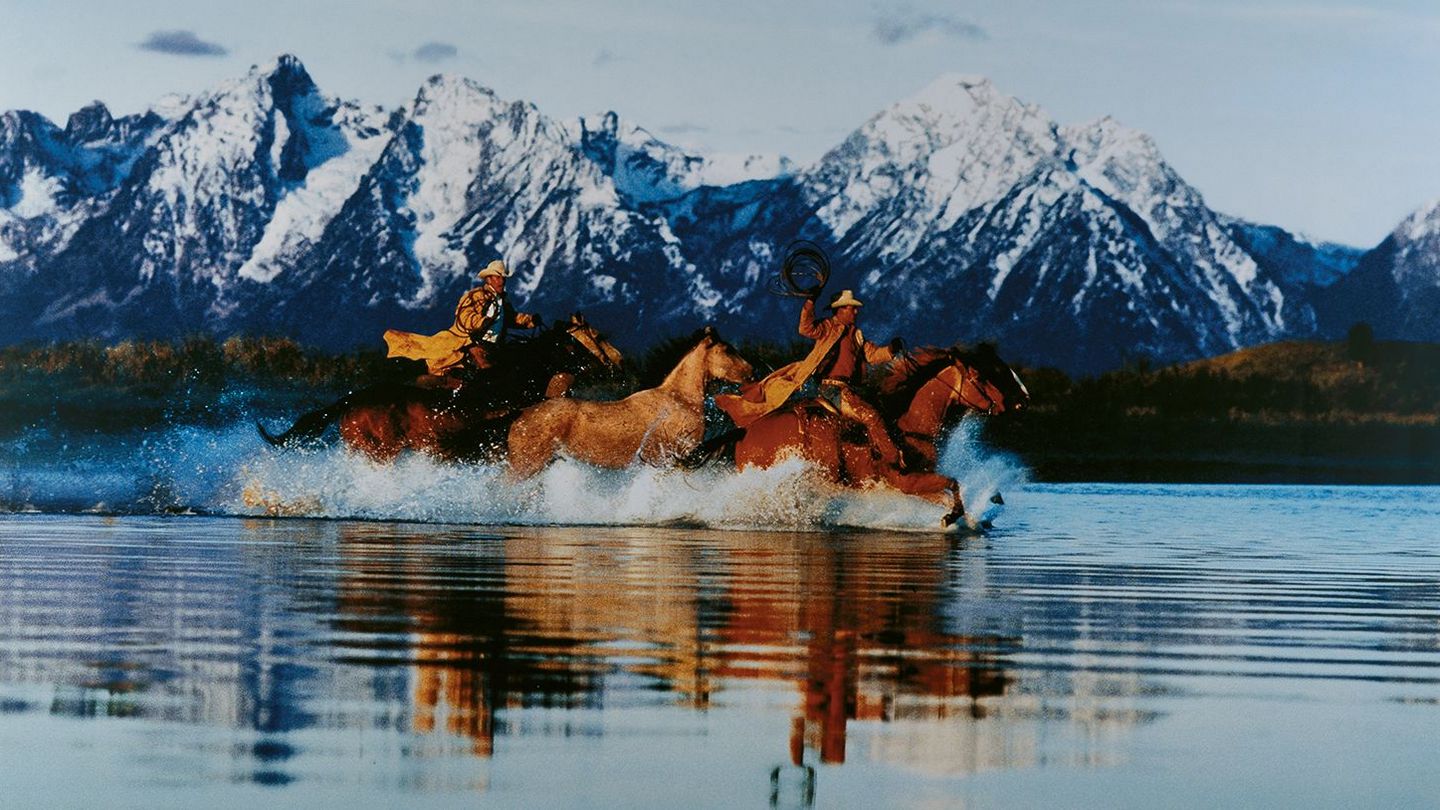 This color photograph shows two cowboys riding three horses through a body of water in front of snowy mountain landscape. Richard Prince, Sammlung Goetz Munich