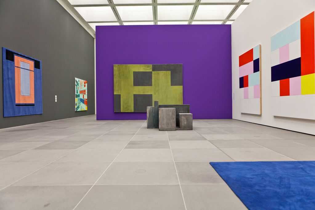 Exhibition view with colourful, minimalist works by the artists Helmut Federle, Peter Fischli/David Weiss, Imi Knoebel, Sarah Morris and Andrea Zittel. These are a wall and floor carpet, four paintings and a floor sculpture in the middle of the room. Helmut Federle, Peter Fischli/David Weiss, Imi Knoebel, Sarah Morris and Andrea Zittel, Sammlung Goetz Munich