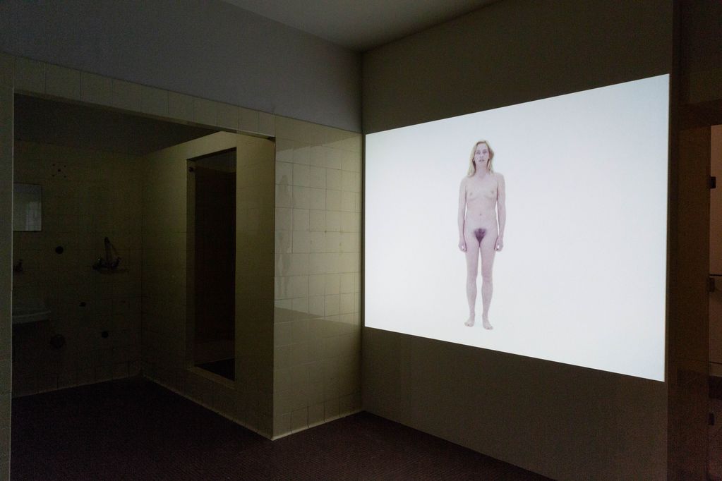 View of the video installation in a room with yellow tiles. The still shows a naked, blonde woman standing in front of a white background. Sam Taylor-Johnson, Sammlung Goetz Munich