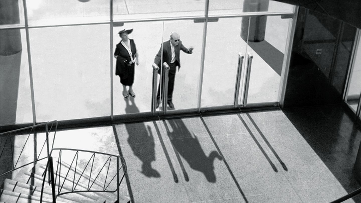 Video Still in schwarzweiß, auf dem ein Mann und eine Frau zu sehen sind, die von einer Wendeltreppe im Inneren eines Gebäudes aus gesehen vor dem verglasten Eingang und in den Raum schauen. David Claerbout, Sammlung Goetz München