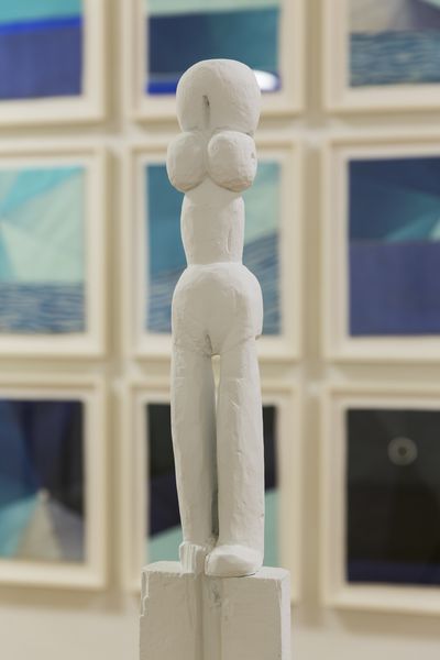 Diese Installationsansicht zeigt eine weiß bemalte, weibliche und langgezogene Skulptur vor einem Hintergrund einer mehrteiligen, zweidimensionalen und bläulichen Arbeit aus Stoff. Louise Bourgeois, Sammlung Goetz