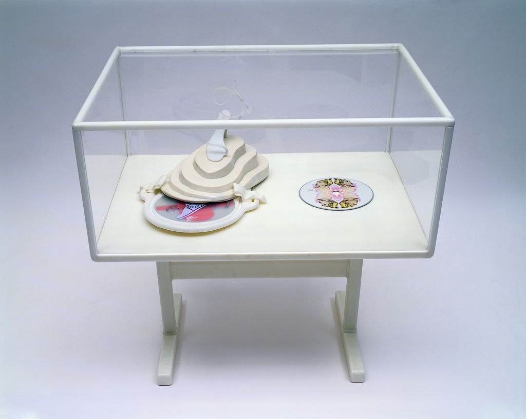 This photograph shows a display case with white edges and a frame in which a white object is placed, on which a white and open high heel is standing. On the right side there is a CREMASTER 1 DVD with the artwork of a person from the film on it.