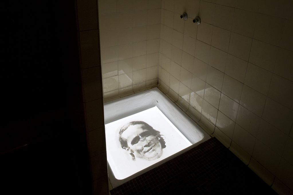 Aufnahme eines dunklen Duschraumes, dessen Duschboden mit einer Videoprojektion bespielt wird. Diese zeigt das Gesicht eines Mannes in schwarzweiß vor hellweißem Hintergrund. Óscar Muñoz, Sammlung Goetz München