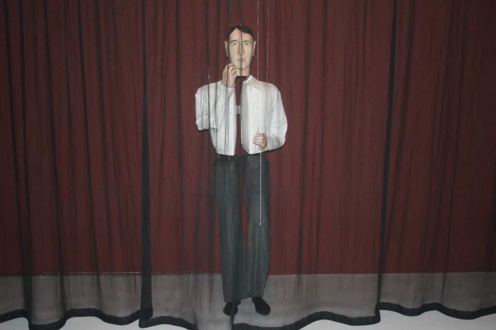 Life-size mannequin of a man in a white chemise and grey suit trousers behind a transparent black curtain. Markus Schinwald, Sammlung Goetz Munich