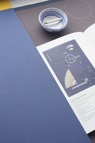 This photograph shows a booklet with a color scheme drawing of Kandinsky, as well as a small white bowl with blue paint and a white colored pencil in it. To the left is clay paper in a warm, bright shade of blue. Kathrin Sonntag, Sammlung Goetz Munich