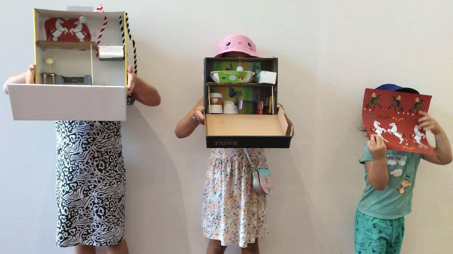 Drei Kinder halten sich selbstgebastelte Puppenhäuser vor das Gesicht