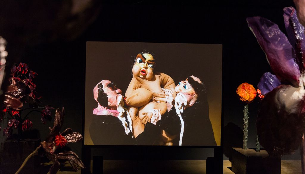 Ausschnitt aus der Rauminstallation "The Experiment", mit Fokus auf einem Bildschirm, dessen Standbild gerade die Figur einer dicken, nackten Frau zeigt, die zwei Mönchsfiguren mit ihren Brüsten säugt. Nathalie Djurberg, Sammlung Goetz München