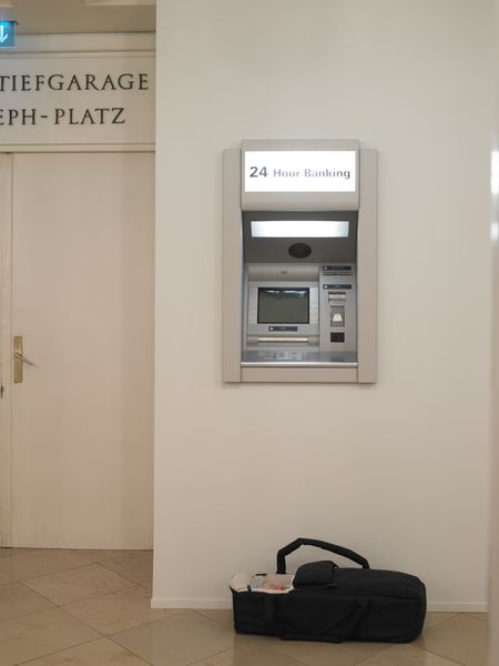 Installationsansicht, die die Arbeit "Modern Moses" zeigt, welche aus einem Bankautomaten besteht, unter dem sich eine Babytragetasche befindet, in dem die Puppe eines Babys liegt. Michael Elmgreen/Ingar Dragset, Sammlung Goetz München