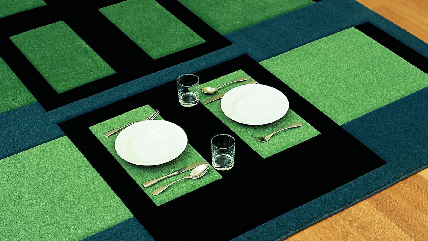 Diese Ausstellungsanischt zeigt einen Teppich der Künstlerin Andrea Zittel, auf dem man (zu Abend) essen kann. Er besteht aus moosgrünen, blauen und schwarzen, rechteckigen Formen, in denen zwei Teller, Bestecke und Gläser gegenüber voneinander aufgestellt sind.