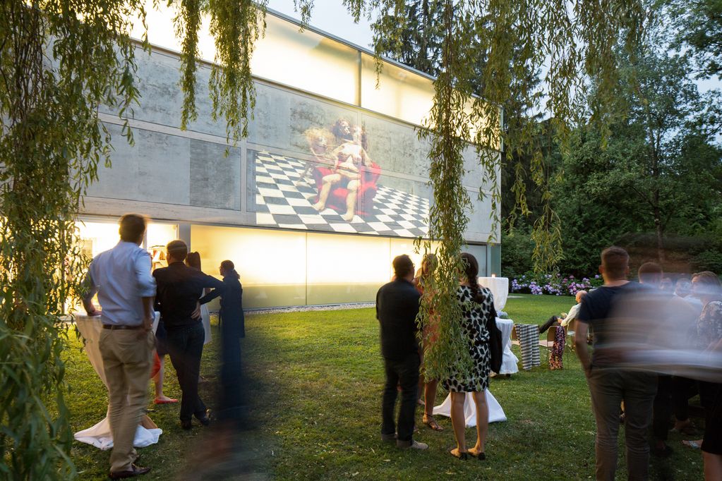 Fotografie eines Open-Air Filmfest der Sammlung Goetz mit Filmen der Künstlerin Nathalie Djurberg. Darauf sind Personen im Garten der Sammlung Goetz stehend zu sehen, sowie das Sammlungsgebäude samt Projektion eines Films auf dessen hölzerne Außenpaneele.