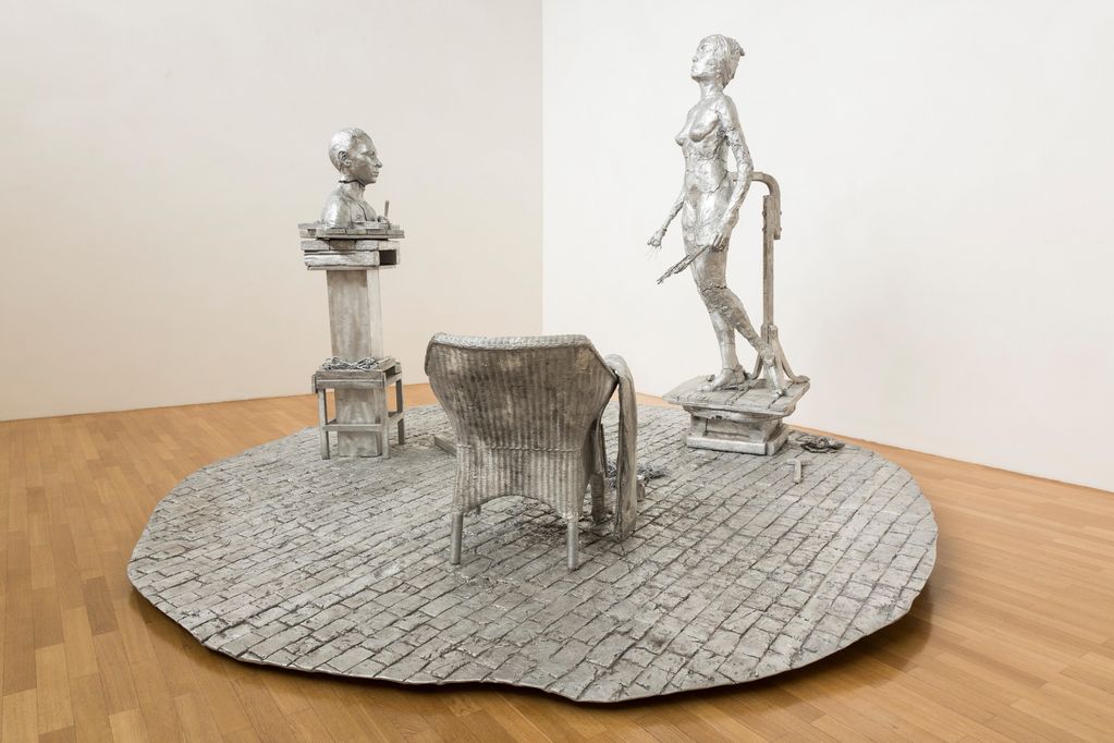 Aluminiumskulptur, bestehend aus einem Steinboden mit einem Korbstuhl darauf sowie einem Sockel mit männlicher Büste und einer nackten Frauenfigur. Paweł Althamer, Sammlung Goetz München