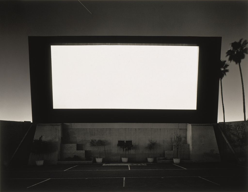 Black and white photograph of an open-air cinema with a white luminous screen and palm trees in the background on the right. Hiroshi Sugimoto, Sammlung Goetz Munich