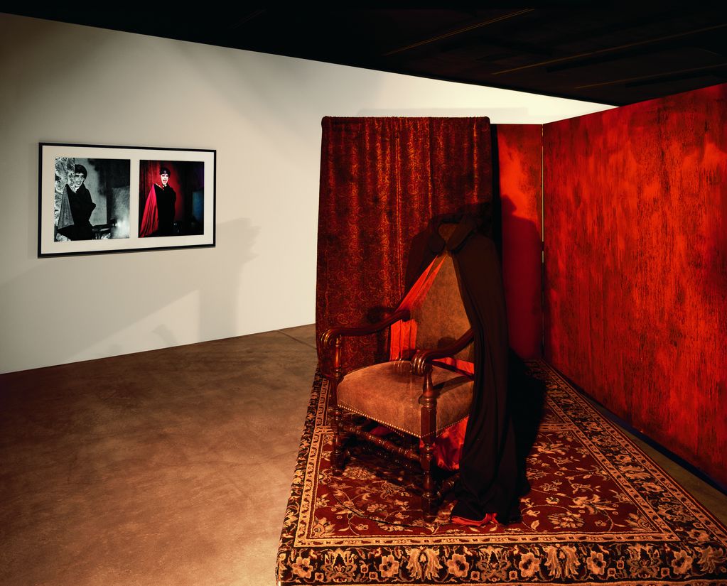 Diese Ausstellungsansicht der Arbeit "Lonely Vampire" zeigt einen altmodischen, gepolsterten Stuhl, über den ein schwarzer Umhang gehängt ist. Dieser steht auf einer erhobenen Fläche, die mit einem rötlichen Perserteppich abgedeckt ist. Dahinter befinden sich zwei Stellwände, die eine halb verdeckt durch einen Brokatvorhang, die restlichen Wände rot angestrichen, die Farbe scheint von den Wänden herabzufließen. Die dahinter befindliche weiße Wand trägt zwei gerahmte Fotografien eines Mannes in einem Vampirkostüm. Mike Kelley, Sammlung Goetz München