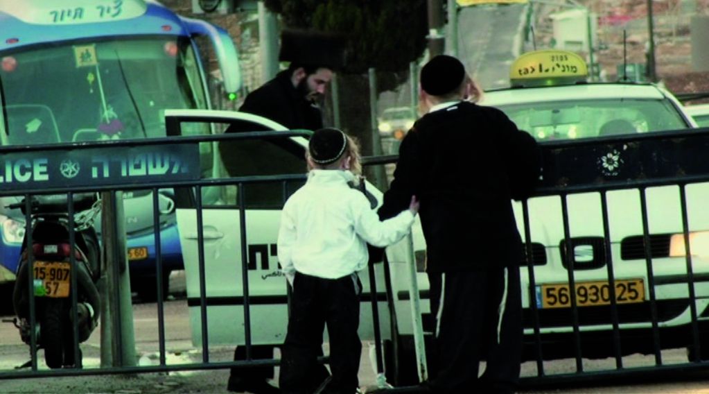 Video Still of two boys in Jewish Orthodox robes and haircuts, with their backs to the viewer and holding hands. They are standing in front of a barrier, behind them is a taxi into which a man in Jewish Orthodox robe and haircut is getting. Nira Pereg, Sammlung Goetz Munich