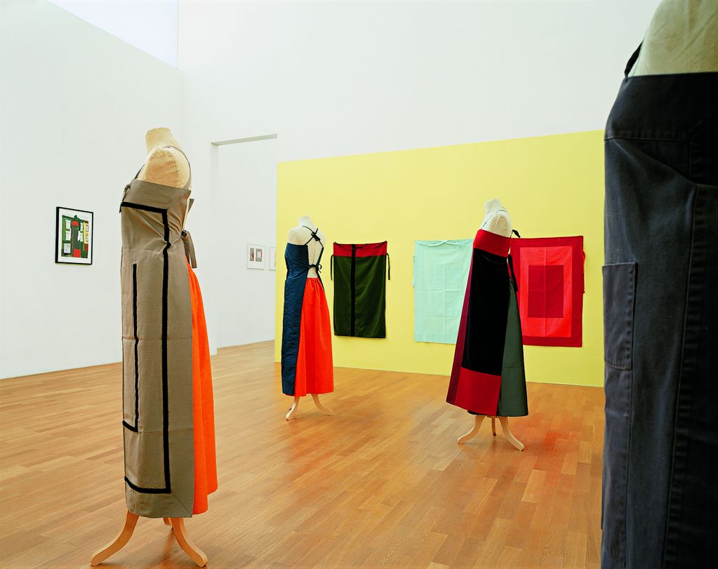 Hier ist eine Ausstellungsansicht mit textilen Wandobjekten und Schürzenkleidern, die auf vier gleichen Schneiderpuppen drapiert sind, zu sehen. An der linken Wand befindet sich ein Entwurf zu einem der ausgestellten Schürzenkleider. 