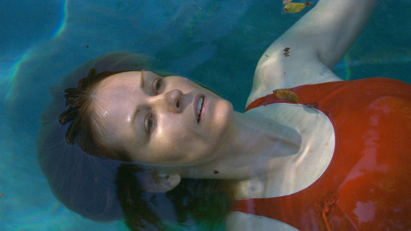 Video Still of a woman who seems to float in the turquoise water of a swimming pool. She has white skin, brunette hair and fine features and wears a tomato-red swimming costume. Teresa Hubbard/Alexander Birchler, Sammlung Goetz Munich