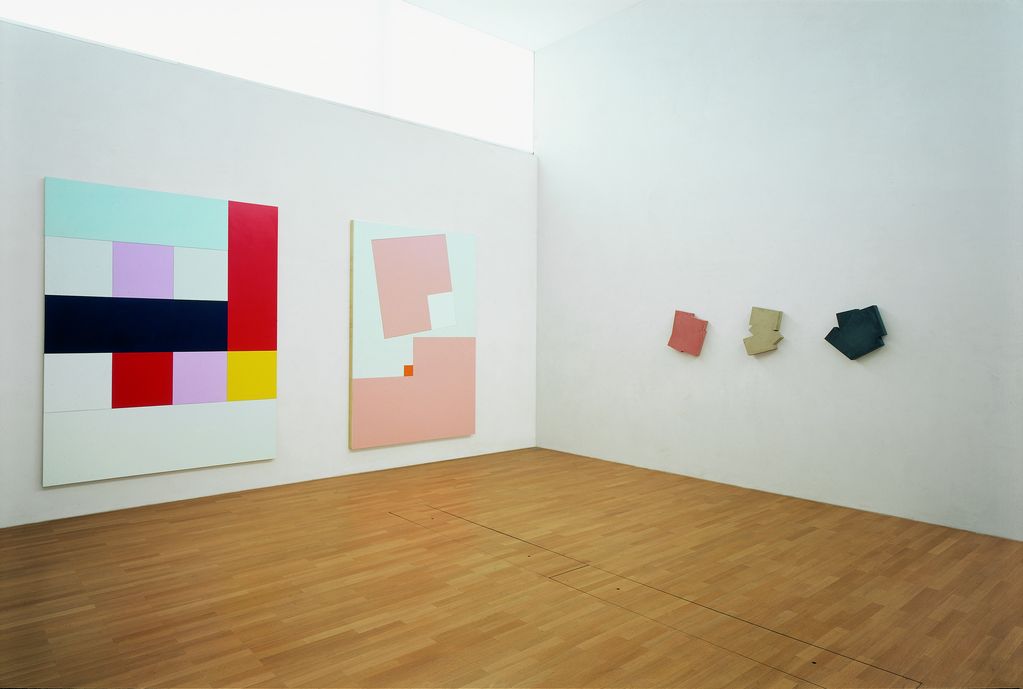 The installation photograph shows three works by the artist Imi Knoebel. Two large-format paintings hang on the left wall, one a little more colourful, the other in white and pink. On the right wall hang the Betonis, monochrome painted objects in a playful form made of concrete.