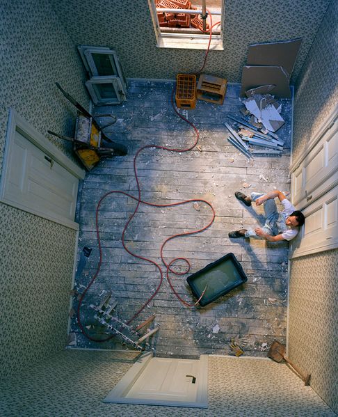 This color photograph shows a housing space seen from a bird's eye view. In it, a young man in dungarees sits leaning against one of the three doors; around him are objects and materials for renovation. Teresa Hubbard/Alexander Birchler, Sammlung Goetz Munich