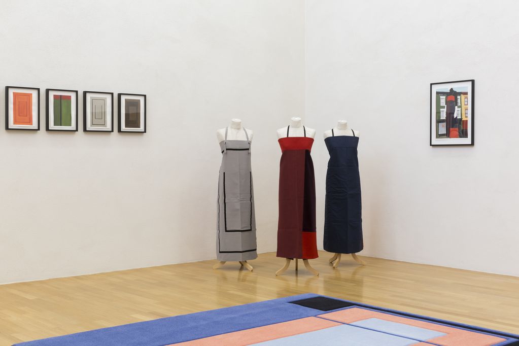 Exhibition space with small-format framed works on one wall, three headless mannequins with minimalist long aprons, the matching sketch hanging on the other wall and a carpet on the floor. Andrea Zittel, Sammlung Goetz Munich