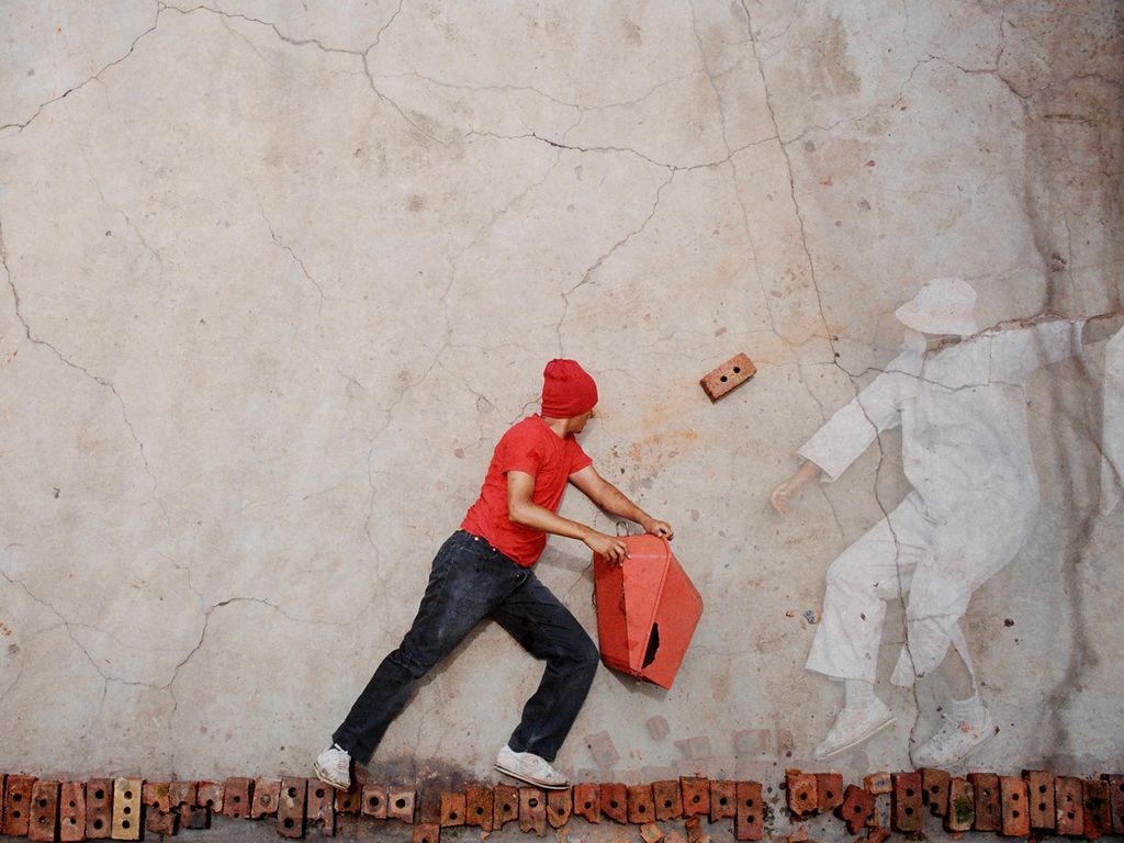 Dieses Video Still zeigt einen Mann mit rotem T-Shirt sowie Mütze und blauen Jeans auf einer Ziegelsteinmauer laufen, während ein Ziegelstein auf ihn zuzufliegen scheint. Rechts neben ihm befindet sich eine nicht ganz deutliche menschliche Gestalt in einem weißen Schutzanzug, Gesichtsmaske und Hut. Robin Rhode, Sammlung Goetz München