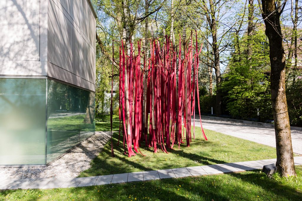 Photograph of an installation by the artist Ulrike Ottinger in the garden of the Sammlung Goetz. These are thin, long and variously red strips of fabric that seem to hang down from the thin white trunks of the birch trees. Ulrike Ottinger, Sammlung Goetz Munich