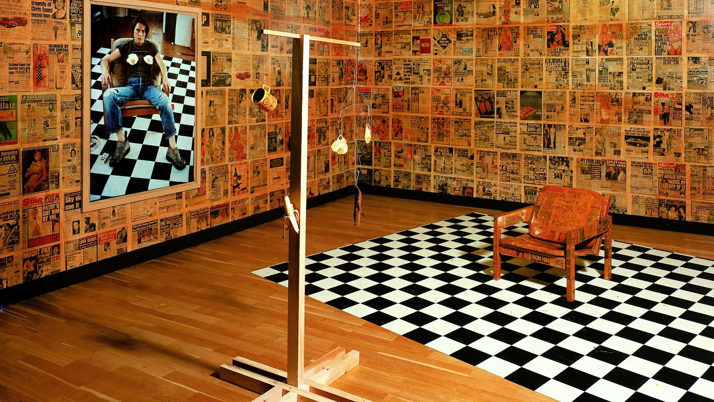 Here you can see a fully decorated room of the artist Sarah Lucas. The walls are covered with posters of newspapers, on the left wall hangs a photographic self-portrait with fried eggs placed on her chest. In the self-portrait she is sitting on a chair which is standing on a chessboard-like background. These two objects can also be found in the room, the chair has now also been provided with newspaper cuttings. In front of it there is a kind of mobile with fried eggs, cup, banana and old banana skin.