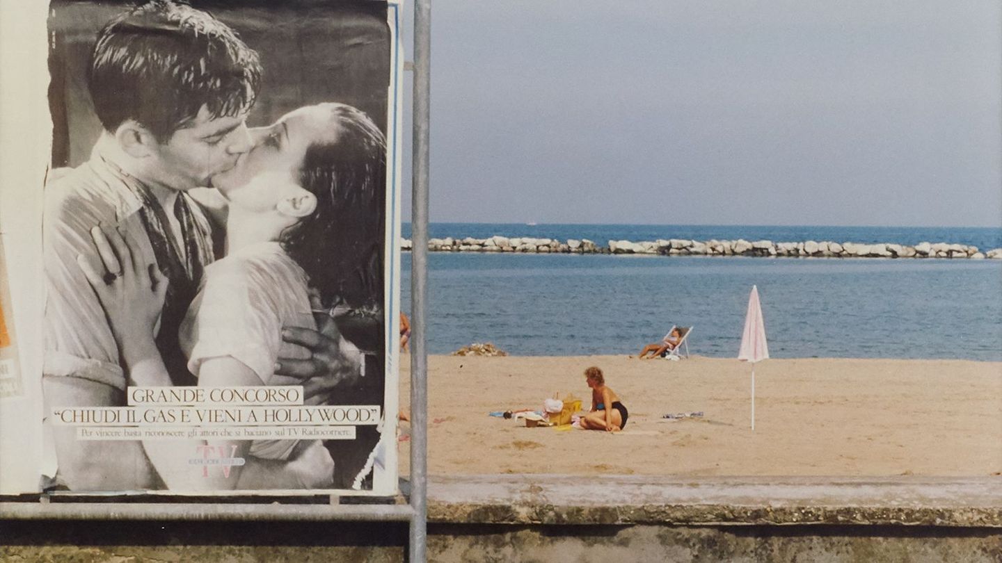 Fotografie einer Strandszenerie mit einem Schwarzweiß-Plakat eines sich küssenden Paares davor. Luigi Ghirri, Sammlung Goetz München