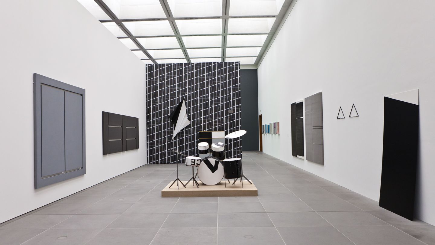 Exhibition view with works exclusively in the colours black, white and grey. These are painted or printed canvases, a wallpaper, a drum set and wall objects. Martin Boyce/Alan Charlton/Wade Guyton/Reiner Ruthenbeck/Michael Sailstorfer, Sammlung Goetz Munich
