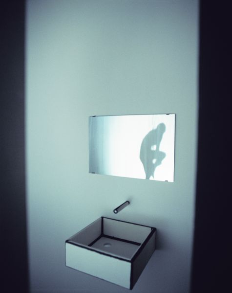 Photograph of a small room installation consisting of a rectangular mirror with a shadow of a female person behind a curtain in it and a simple water tap plus washbasin made of paper. Zilla Leutenegger, Sammlung Goetz Munich