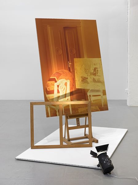 Here we can see an installation consisting of an easel with a large-format, sepia-coloured photograph on it, a stretcher frame leaning against it and a canvas with a white surface below it. The installation is illuminated by a spotlight on the floor.
