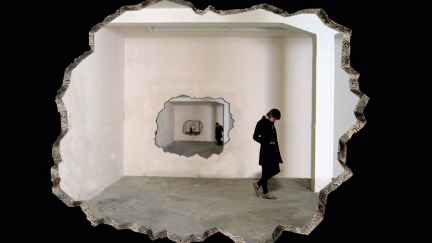 Video projection showing a hole in a wall with a person dressed in black standing behind it. Behind the person is another hole in a wall with a person behind it. This situation seems to repeat itself endlessly. Zilla Leutenegger, Sammlung Goetz Munich