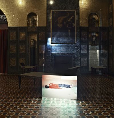 Installation view of the work "Sleepers" by Francis Alÿs in the Villa Stuck. The projection of a still image of a man sleeping in the street is shown. The photograph shows both the projector and the projection as well as the luxurious salon of the Villa Stuck. Francis Alÿs, Sammlung Goetz Munich