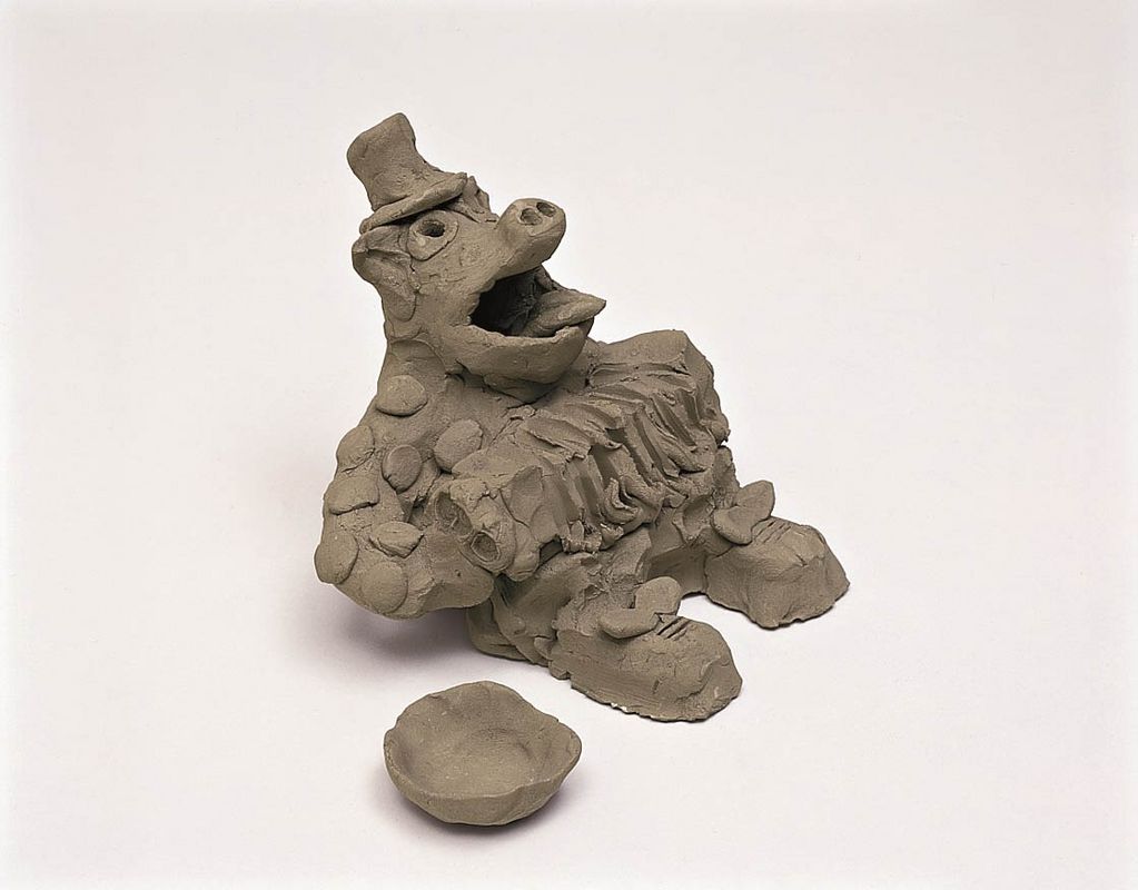 Hier ist eine Skulptur aus grauem Ton zu sehen. Sie stellt ein Tier mit Schweineschnauze und herabhängenden Ohren dar, das einen Hut, ein gepunktetes Oberteil, Hose und Schuhe trägt. Es spielt eine Ziehharmonika, hat den Mund weit geöffnet und eine Schale neben sich aufgestellt.