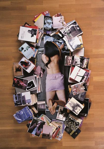 This is a photograph of a reading woman lying on her stomach, seen from a bird's eye view, surrounded by stacks of books. She has long, dark hair and is undressed except for a short cotton skirt. The photograph is a work by the artist Rosemarie Trockel and consists of a life-size photograph of the woman with her head cut out and raised. The books around the woman are real, however. 