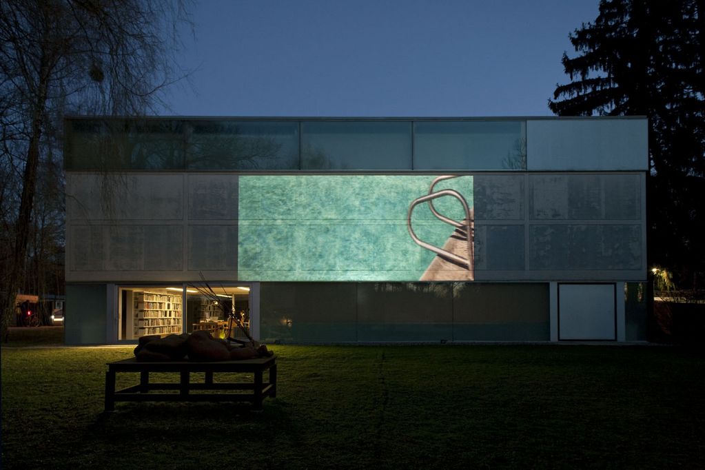 Photo of the Sammlung Goetz at dusk, with a sculpture in front of the brightly lit entrance area. A video by Doug Aitken is projected onto the wooden panels of the collection building, just showing the entrance of a turquoise blue swimming pool. Doug Aitken, Sammlung Goetz Munich