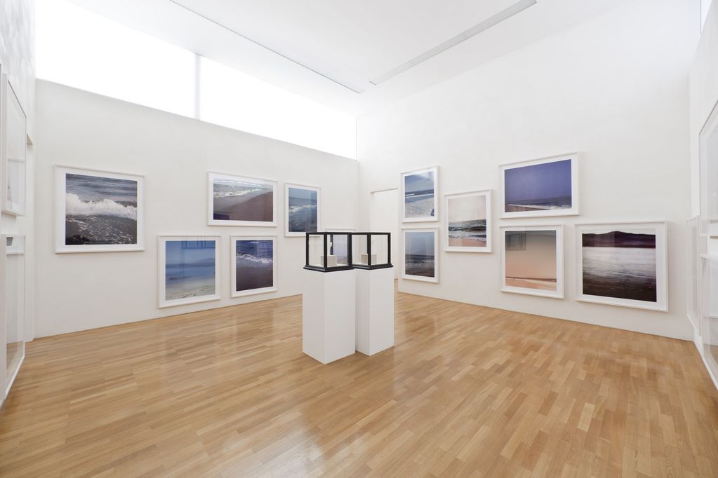 Exhibition view with framed photographs of sea hitting the sand, which are hung next to and above each other. In the center of the room are two pedestals with glass boxes on them, in which two white cubes lie. Paul Pfeiffer, Sammlung Goetz Munich