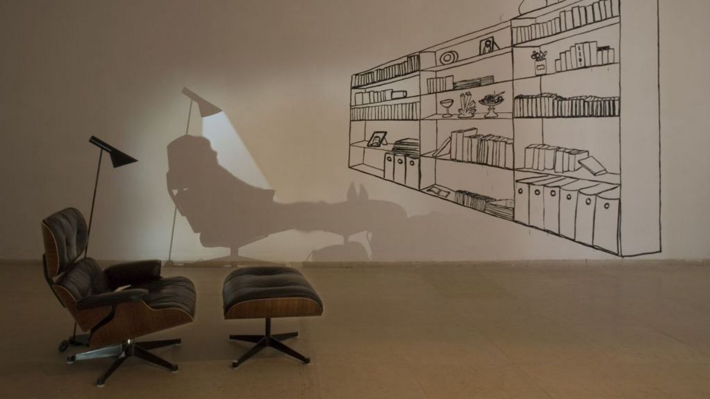Installation view showing a black upholstered armchair with an ottoman, a floor lamp provides light in the otherwise darkened room. To the right in front of it is a drawn shelf, with books, folders and vessels inside. Behind the armchair against the wall, a shadow of the armchair and the floor lamp can be made out; in addition, a person appears to be lying on the armchair and ottoman. Zilla Leutenegger, Sammlung Goetz Munich