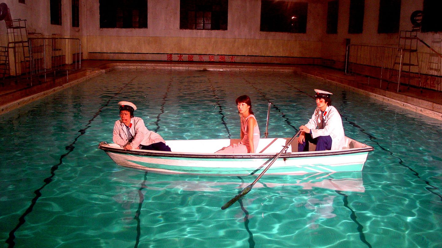 Auf dem Video Still sind drei asiatisch aussehende Personen in einem Boot sitzend zu sehen. Das Boot selbst befindet sich auf dem türkisblauen Wasser eines Schwimmbeckens in einem Schwimmbad. Yang Fudong, Sammlung Goetz München