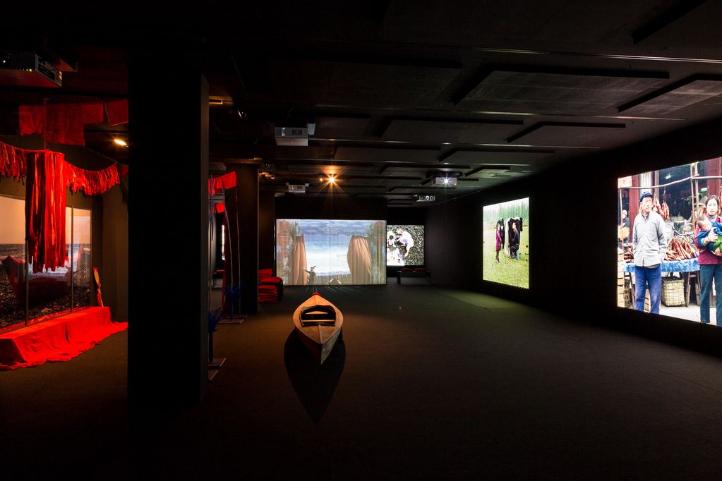 Exhibition view of the work "Floating Food", consisting of several video projections, a small wooden boat in the middle of the room, a photograph as a triptych as well as several red fabric panels serving as a cover or decoration. Ulrike Ottinger, Sammlung Goetz Munich