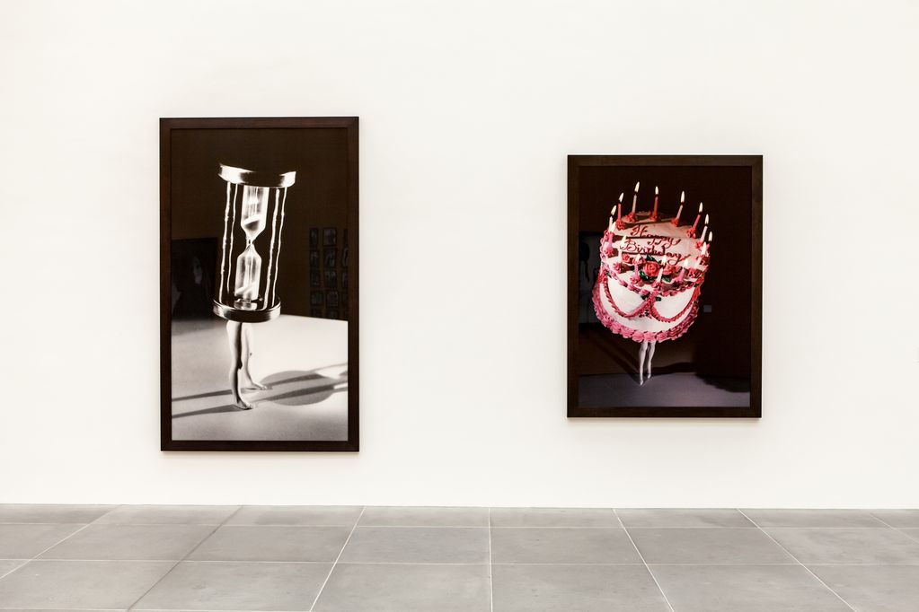Exhibition view with two photographic works by the artist Laurie Simmons. On the left, a black and white photograph of modelled women's legs with an hourglass as their torso is hung on the white wall. To the right is a colour photograph of modelled women's legs with a birthday cake as their upper body. Sammlung Goetz Munich