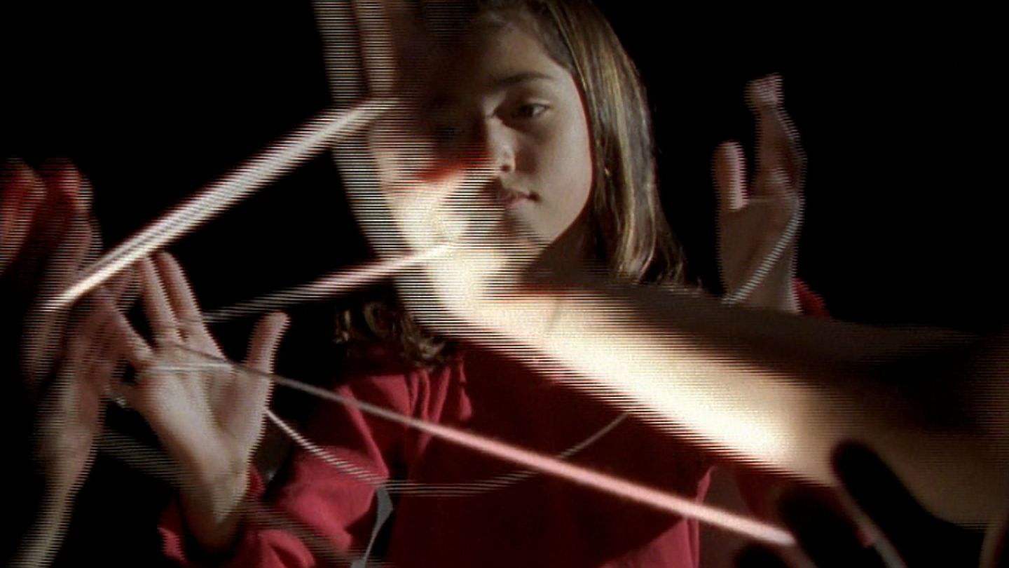 Dieses Filmstill zeigt ein junges Mädchen mit dunklen Haaren in einem dunklen Raum, das ein Spiel mit Fäden zwischen den Fingern spielt. Es scheint isoliert von den anderen mitspielenden Kindern zu sein, von denen man nur die erhobenen Hände mit den gleichen Fäden darin sieht. Doug Aitken, Sammlung Goetz München