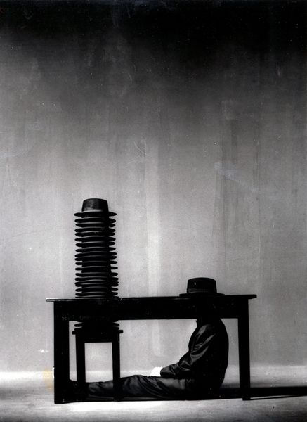 This black and white photograph is kept in a strong light-dark contrast. The table appears almost black in an elongated profile, as does the person sitting on the floor, legs stretched out, face covered by the tabletop. Only a hat peaks out above it. To the left of it, several hats are stacked on top of each other on a stool.