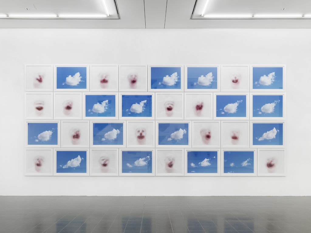 This installation photo shows an arrangement of photographs, alternating between clouds in a rich, blue sky and blurred clown faces. Roni Horn, Sammlung Goetz München