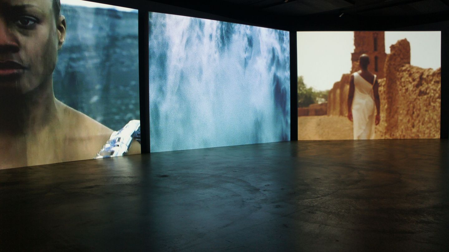 Installation view with three large video projections in a dark room. The first still image of a projection shows half of a young black woman's face in close-up. The second projection depicts falling water and the third shows a black woman in rear view in a white, off-the-shoulder, close-fitting dress in a desert area with mud buildings. Isaac Julien, Sammlung Goetz Munich