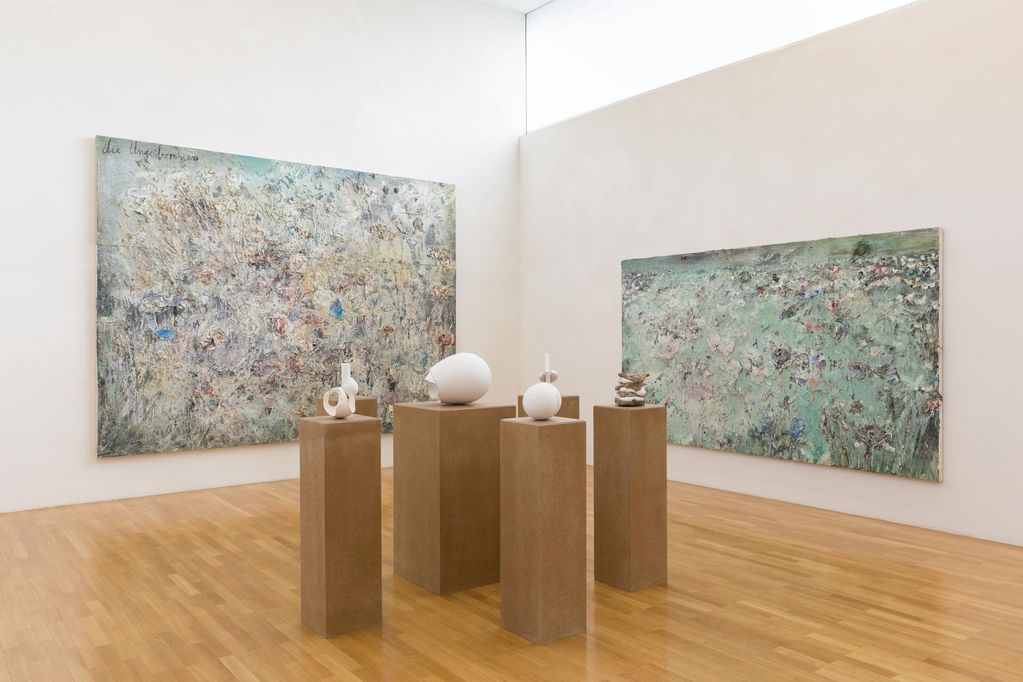 Exhibition view with two abstract and large-format paintings by the artist Anselm Kiefer, in delicate shades of turquoise and brown. In front of them, on light brown plinths, are white plaster works by the artist Maria Bartuszova that resemble organic forms. Maria Bartuszova/Anselm Kiefer, Sammlung Goetz Munich