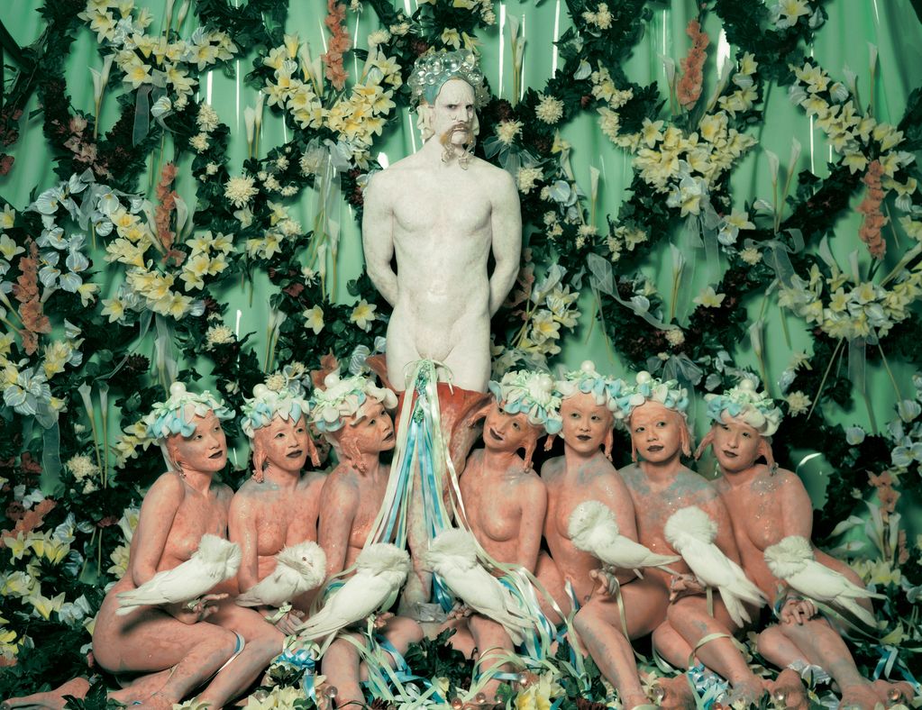 Here you can see a production still from the Cremaster series of the artist Matthew Barney. In front of a flower wall stands a man, painted white and looking like a faun. In front of him sit seven undressed women with strange looking ears and headdresses. They each hold a rare white pigeon on their hand.