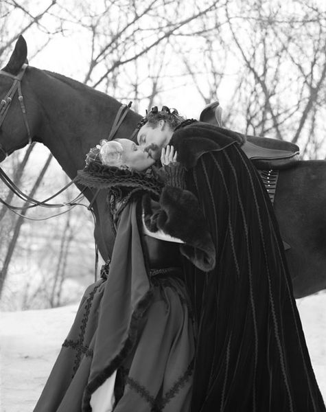 This black and white photograph shows a woman and a man in wintery renaissance clothing, standing in front of a horse and kissing passionately.