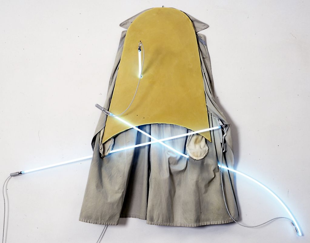 Wall object consisting of a trench coat partially covered by a wooden board covered with yellow wax. Three thin neon tubes glowing light blue are inserted through both. Mario Merz, Sammlung Goetz Munich